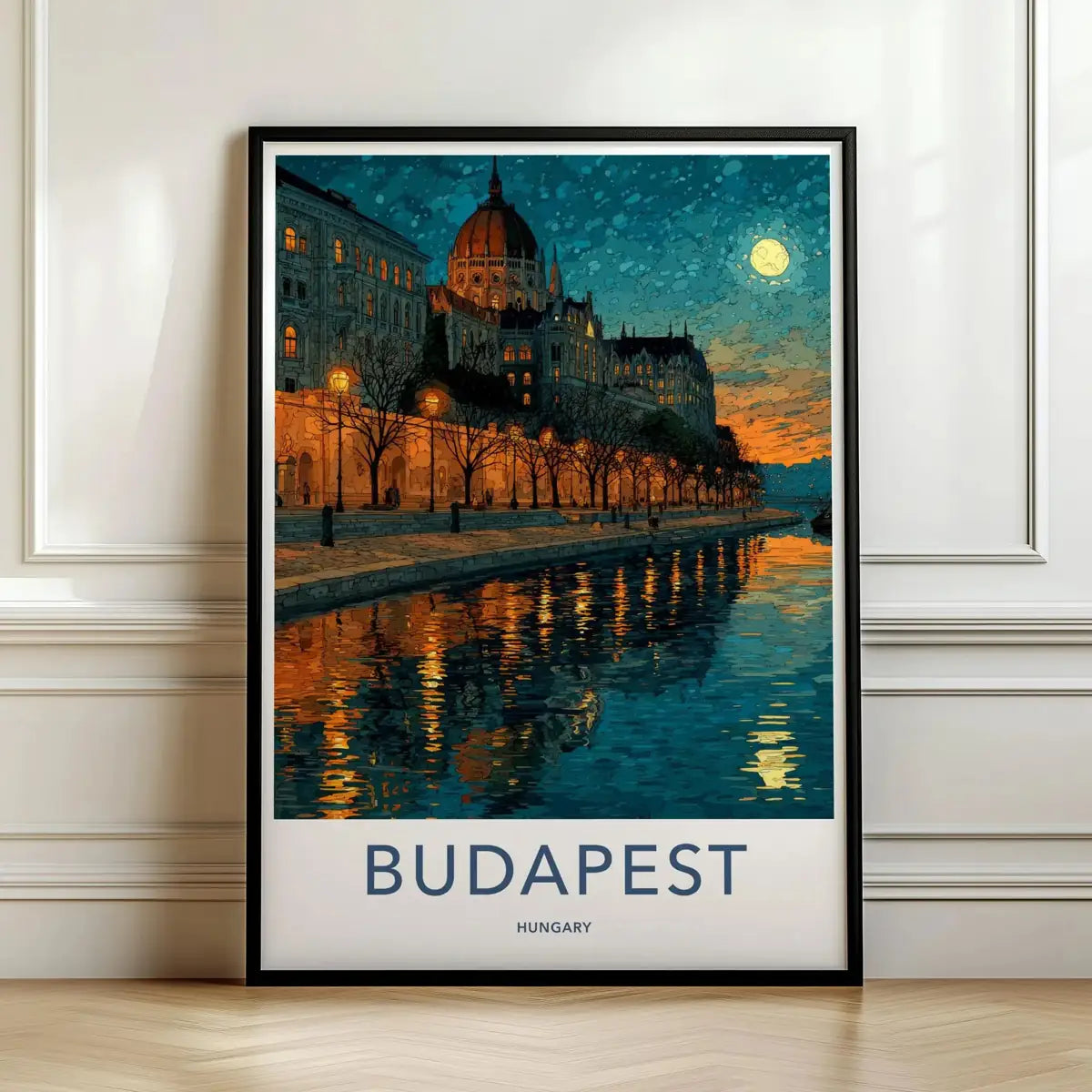 Framed travel poster of Budapest's Parliament building illuminated at night along the river leaning against a white wall.