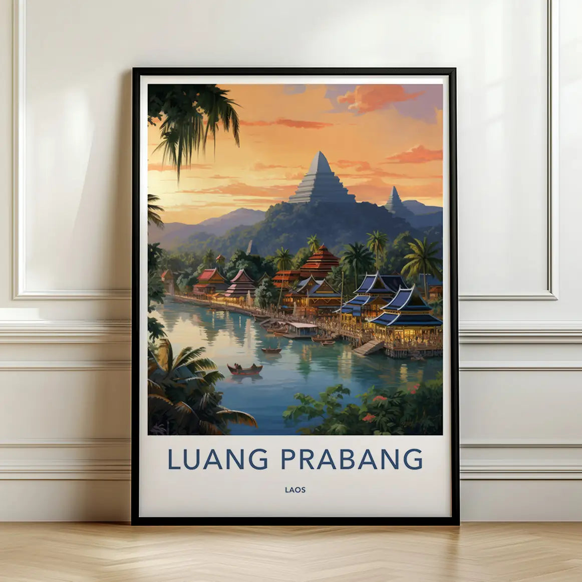 Framed travel poster of Luang Prabang's traditional riverside temples and mountain landscape at sunset leaning against a white wall.