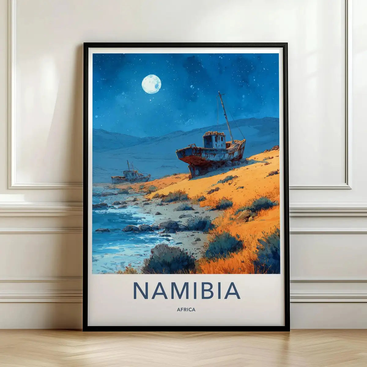 Framed travel poster of a shipwreck on a Namibian beach under a glowing full moon leaning against a white wall.