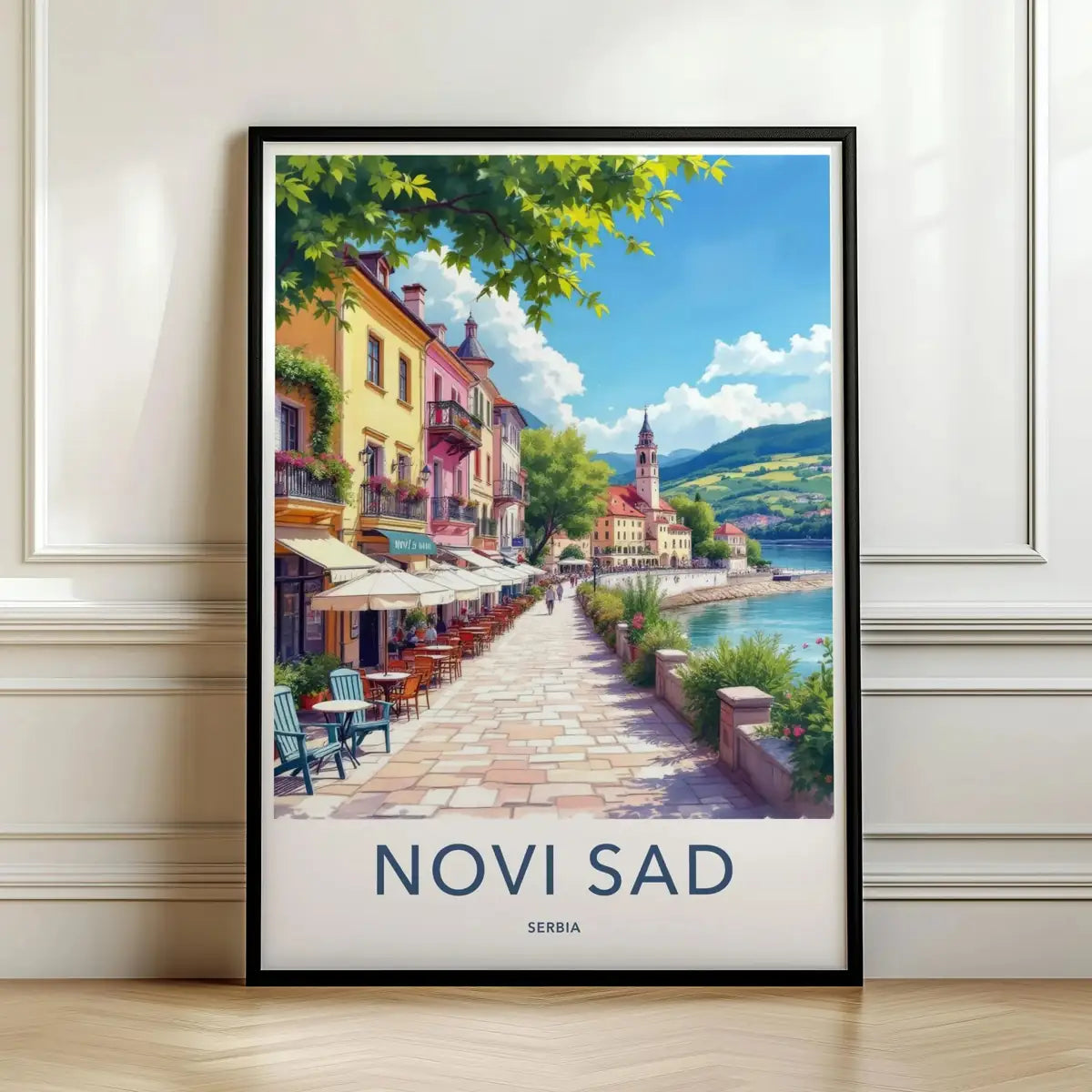 Framed travel poster of Novi Sad's riverside architecture and outdoor cafe leaning against a white wall.