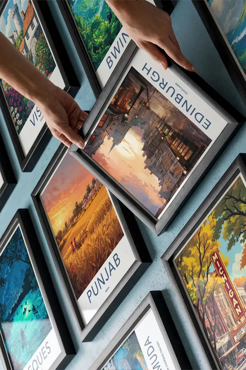 A person's hands are shown holding a framed travel poster of Edinburgh, UK, against a light blue wall displaying other framed posters.