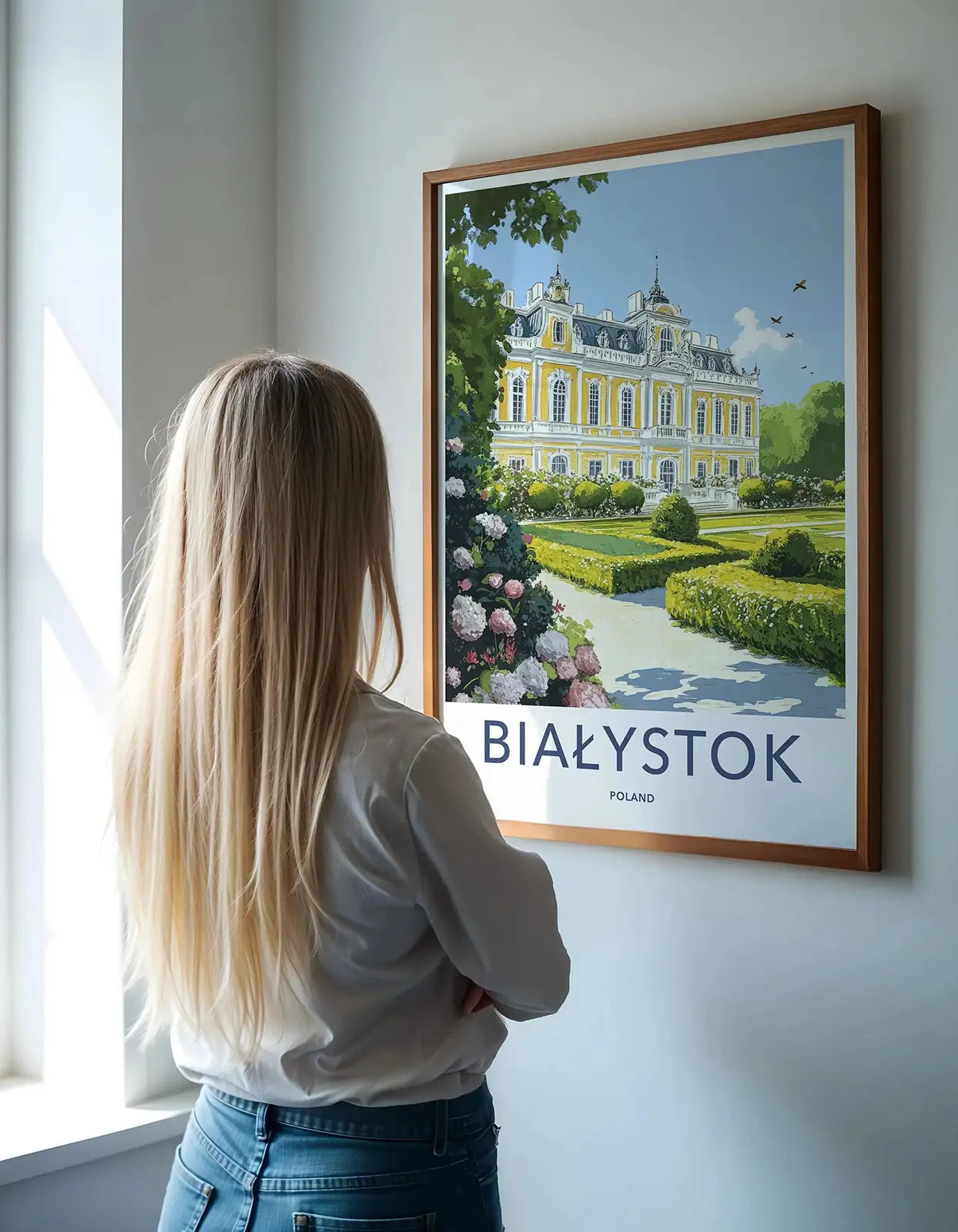 A woman admiring a framed Bialystok Poland travel poster hanging in a bright gallery room.