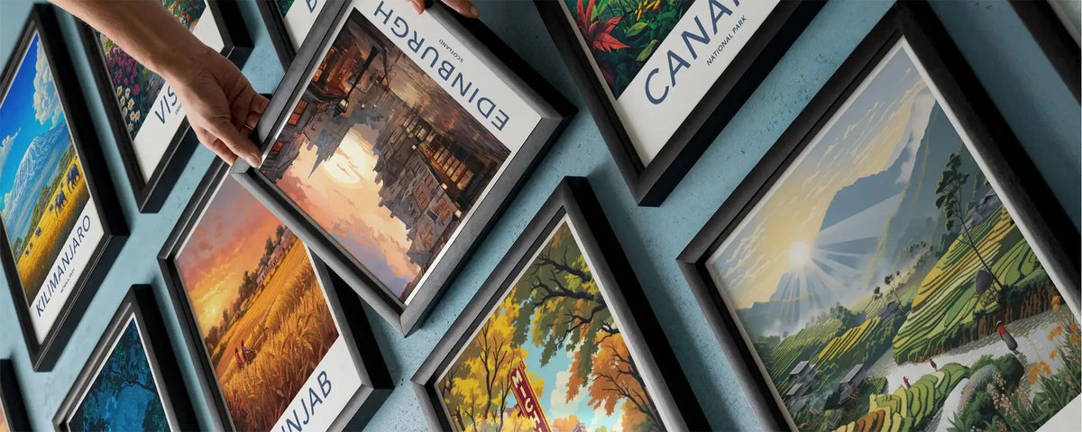 A hand holds a framed landscape art print featuring Edinburgh, Scotland, against a light blue wall with other framed prints of various landscapes and scenes.