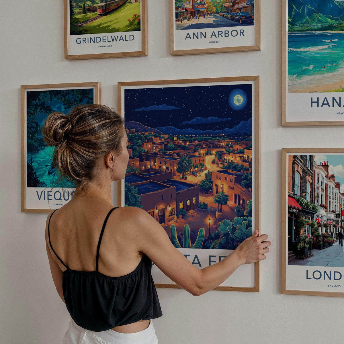 A woman with her hair in a bun is seen from behind, adjusting a framed art print on a white wall. The central framed print shows a colorful night-time illustration of a desert town with cacti and warm glowing buildings under a starry sky.