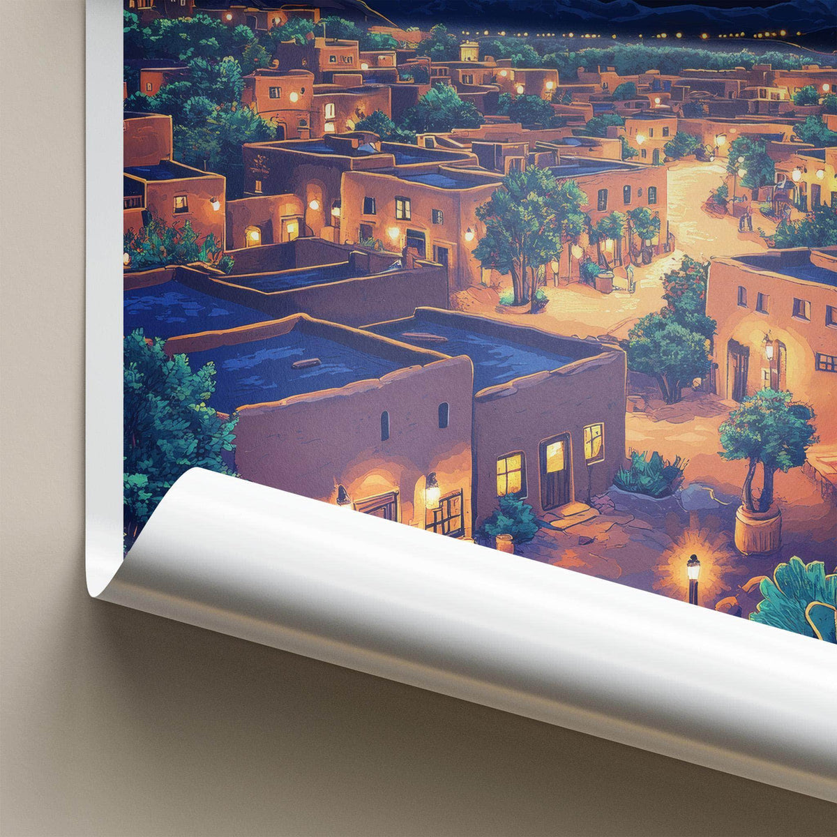 A rolled poster or wall art print is shown against a light beige wall, revealing a colorful illustration of a desert-style village at night with tan adobe buildings, warm glowing windows, green trees, and blue rooftops.